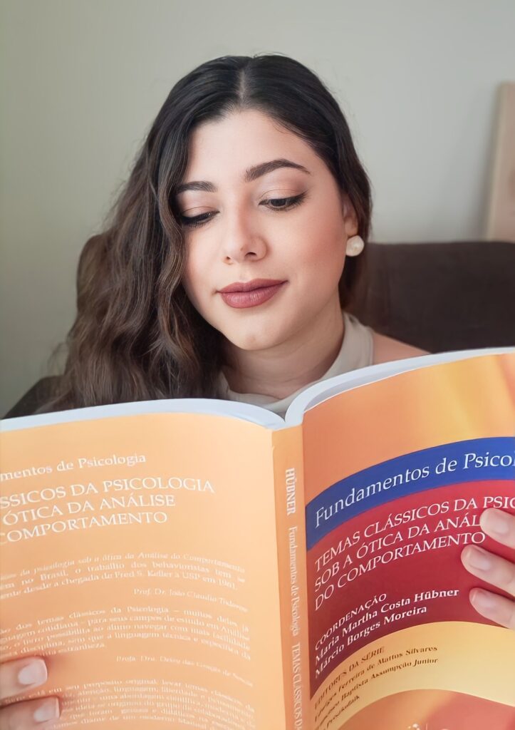 Foto da psicóloga Jéssica Cândido, com um livro de capa alaranjada, sobre Psicologia Comportamental